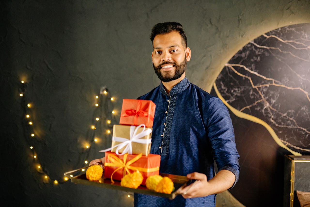 A cheerful man in traditional attire holds a tray with colorful gifts and flowers, perfect for Diwali.